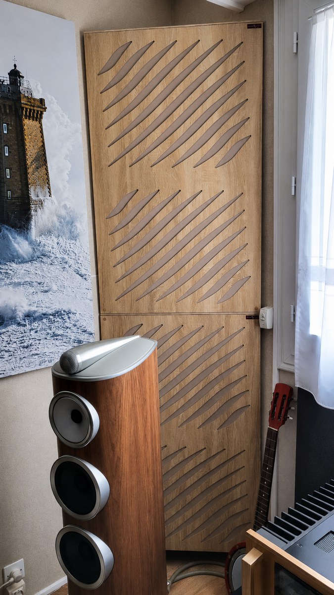 Bass trap angolare con diffusore in finitura legno chiaro, composto da moduli sovrapposti con tagli diagonali, posizionato in un angolo accanto a un diffusore da pavimento e a una chitarra.
