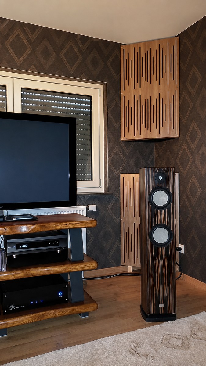 Bass trap angolare con diffusore frontale in finitura legno, installato nell’angolo di una sala d’ascolto accanto a diffusori da pavimento e mobile hi-fi.
