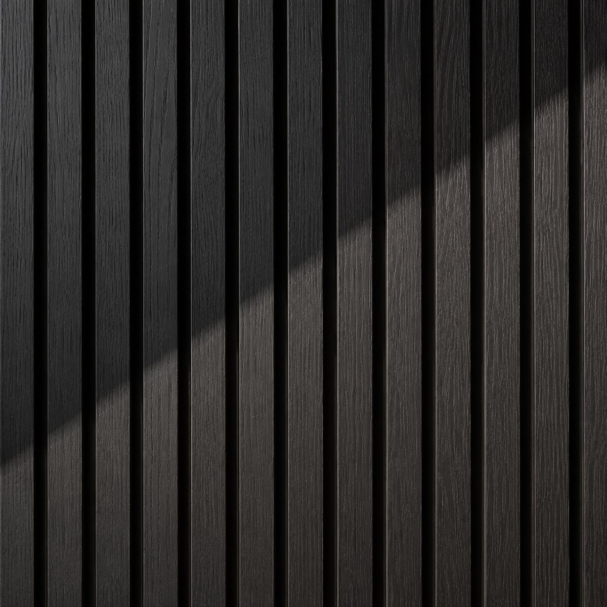 Primo piano di un pannello fonoisolante Freja in legno con lamelle verticali scure e sottili fughe nere, illuminato da una fascia di luce diagonale che mette in risalto la venatura del legno.